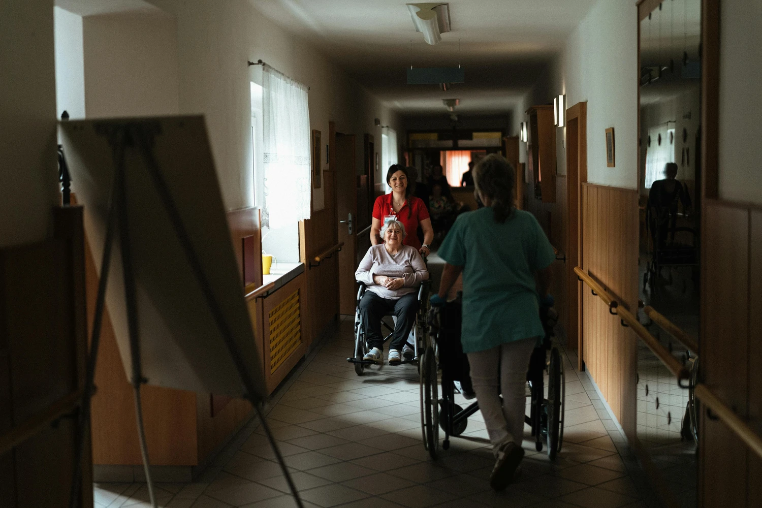 AB Ambulanter Pflegedienst - Traunstein  Ambulanter Betreuungs und Pflegedienst - Eine Pflegekraft hilft einem älteren Bewohner im Rollstuhl auf dem Flur einer Langzeitpflegeeinrichtung.